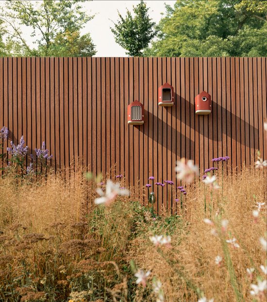 Drie roodbruine insectenhotels hangen aan een bruin rasterhek, omringd door planten.