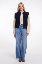 Model draagt een donkerblauwe, fluffy gebreide gilet met ronde hals over een crèmekleurige blouse, wijde jeans en bruine schoenen.