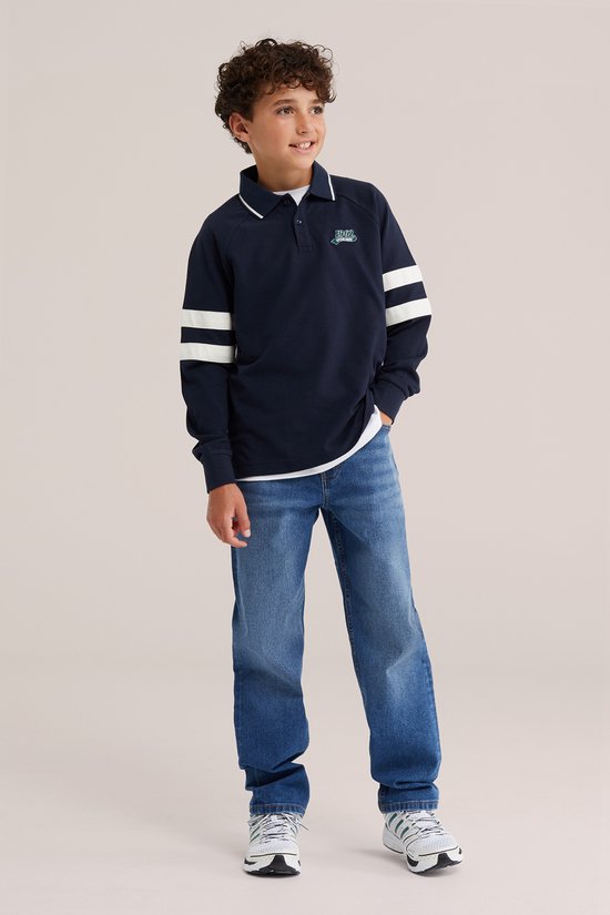 Jongen draagt een blauwe loose fit spijkerbroek, witte sneakers met groene strepen en een donkerblauw poloshirt.