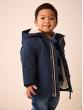 Jongetje lacht, draagt donkerblauwe parka met teddy voering, blauwe trui en spijkerbroek.