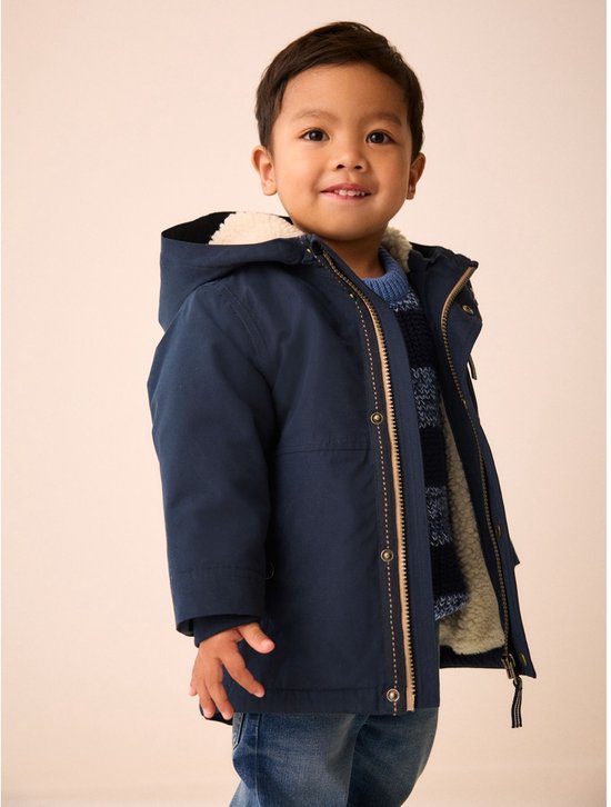 Jongetje lacht, draagt donkerblauwe parka met teddy voering, blauwe trui en spijkerbroek.