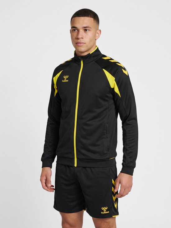 Man draagt zwarte Hummel trainingsjas met gele accenten en bijpassende shorts.