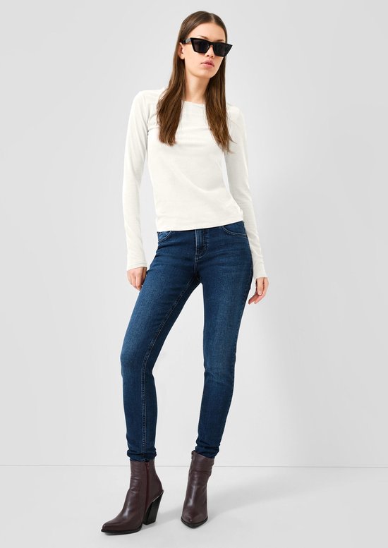Model draagt een wit longsleeve shirt, donkerblauwe jeans, bruine enkellaarsjes en een zonnebril.