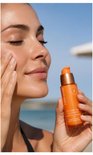 Vrouw met gebruinde huid brengt Lancaster Golden Tan Maximizer serum aan, met strand op de achtergrond.