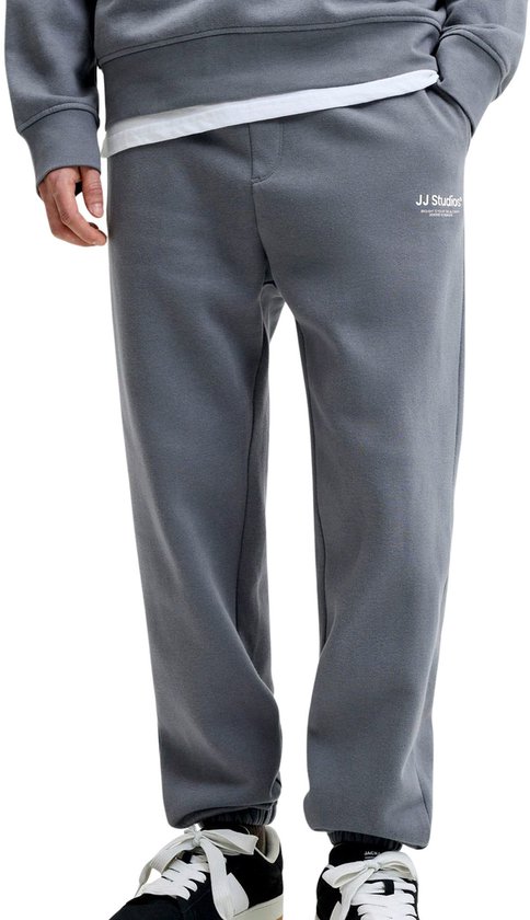 Man in grijze joggingbroek en bijpassende sweater. De broek heeft 'JJ Studios' op de zijkant.