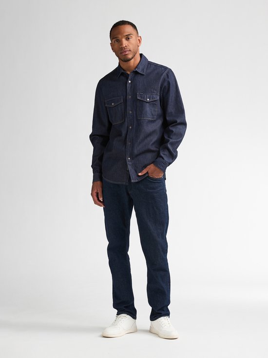 Man in donkerblauw denim overhemd met lange mouwen, twee borstzakken en jeans.