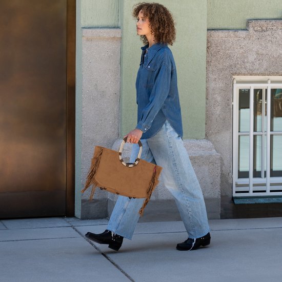 Model loopt met een bruine suède tas met franjes en gouden handvatten. Ze draagt een spijkerblouse en jeans.