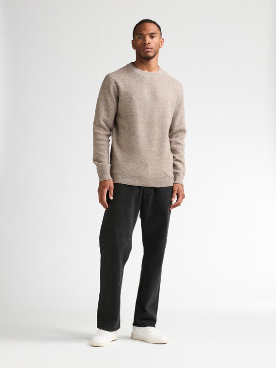 Man in een beige gebreide trui en zwarte loose fit jeans, staand.