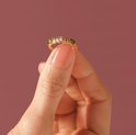 Gouden ring met vijf hartvormige stenen in groen, lichtroze en wit, vastgehouden tussen duim en wijsvinger.