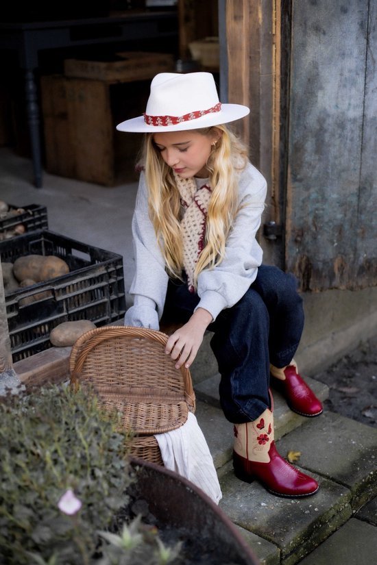 Meisje met witte hoed en rode cowboylaarzen met vlinderborduursel zit op een trap met een rieten mand.