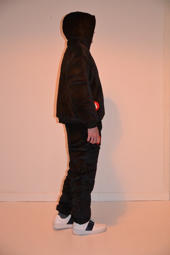 Zijaanzicht van een man in een oversized zwart trainingspak met capuchon. De capuchon is omgeslagen en toont witte voering. Op de linkerbroekspijp is een rood S-logo zichtbaar. Hij draagt witte sneakers.