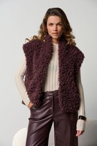 Model draagt een donkerbruine, fluffy gilet met een brede kraag over een crème coltrui en bruine leren broek.