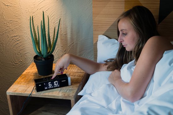 Vrouw raakt een zwarte Lenco wekkerradio aan met tijd '06:10' en zender 'BBC WORLD SERVICE' op een houten nachtkastje.