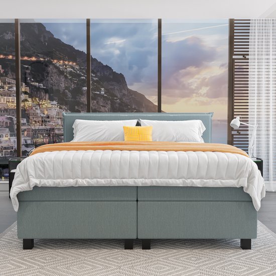 Lichtblauwe boxspring met opgemaakt bed en uitzicht op een bergachtige kuststad.