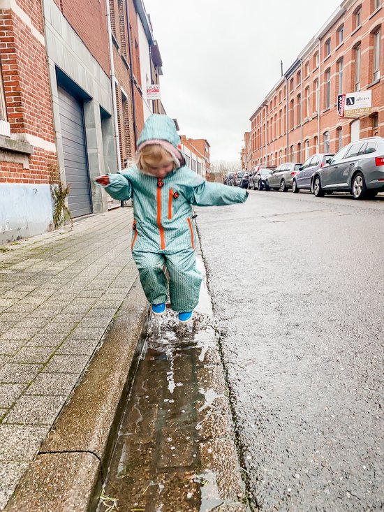 Kind in een lichtgroen regenpak met visgraatpatroon en oranje ritsen, springend in een plas op straat.