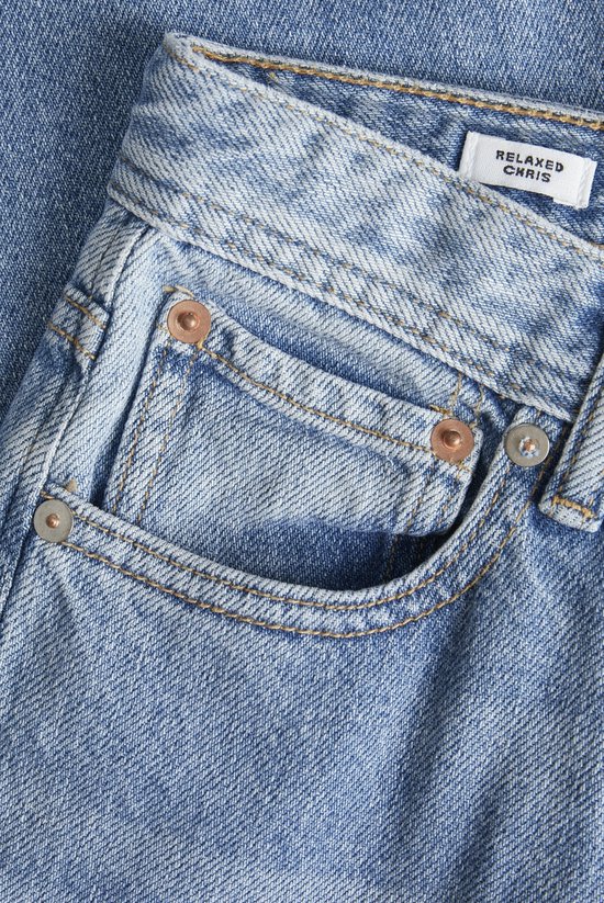 Detailopname van een lichtblauwe spijkerbroek met koperen en zilveren knopen en een 'RELAXED CHRIS' label.