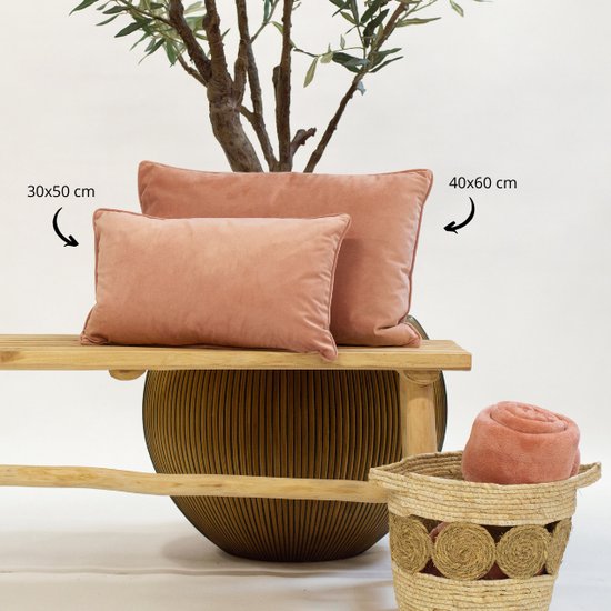 Deux coussins décoratifs en velours rose sur un banc en bois, à côté d'un panier contenant une couverture roulée.