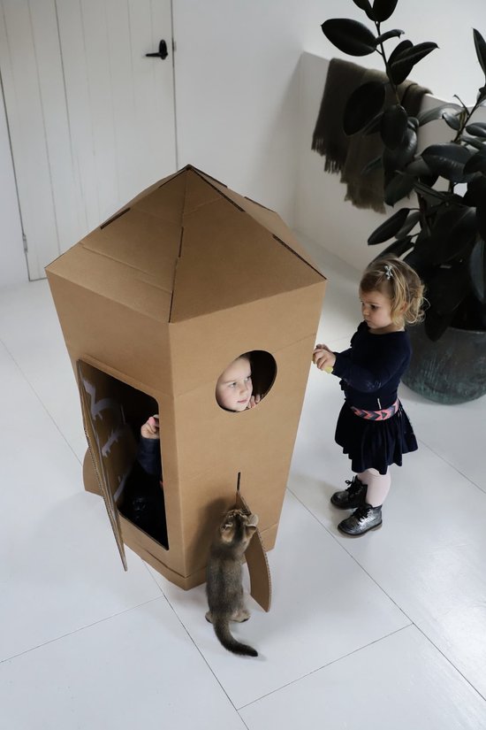 Kartonnen raket met open deur. Een jong meisje kijkt naar buiten vanuit een raam. Een ander meisje en een kat staan naast de raket.