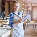 Vrolijke vrouw in schort en geruite blouse met tablet in een werkplaats.