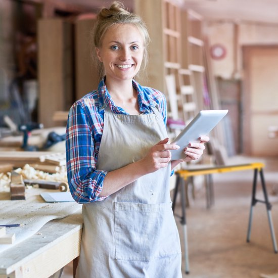 Vrolijke vrouw in schort en geruite blouse met tablet in een werkplaats.