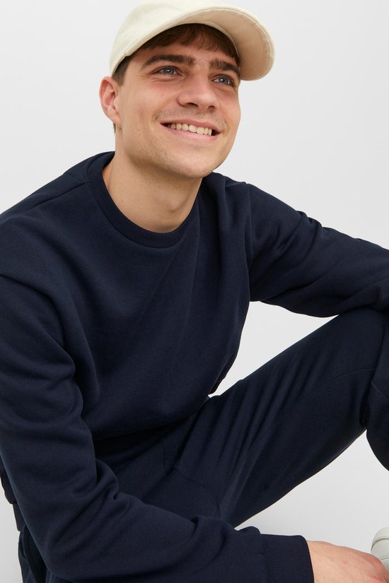Model in een donkerblauwe sweater en joggingbroek, met een beige pet.