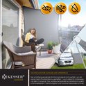 Een vrouw zit op een stoel op een balkon onder een grijs schaduwdoek, met een camper zichtbaar in de achtertuin.
