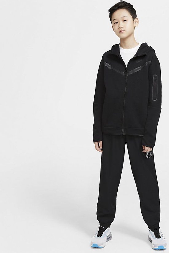 Jongen staat in een compleet zwarte Nike Tech Fleece outfit met vest en broek.
