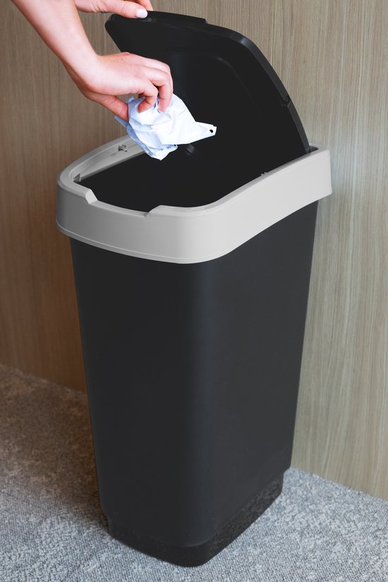 Een hand opent het deksel van een zwarte prullenbak met grijze rand, om een stuk papier weg te gooien.