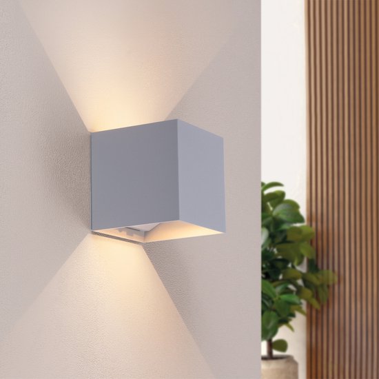 Applique murale Ledvion grise projetant une lumière blanc chaud vers le haut et vers le bas, sur un mur à côté d'une plante.