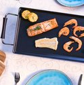 Gegrilde zalm, kabeljauw, maïs en garnalen op een zwarte bakplaat met een klassiek tafelkleed.