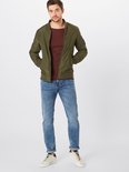 Man in een lichtblauwe jeans, groen bomberjack en bordeaux T-shirt, staand met sneakers.