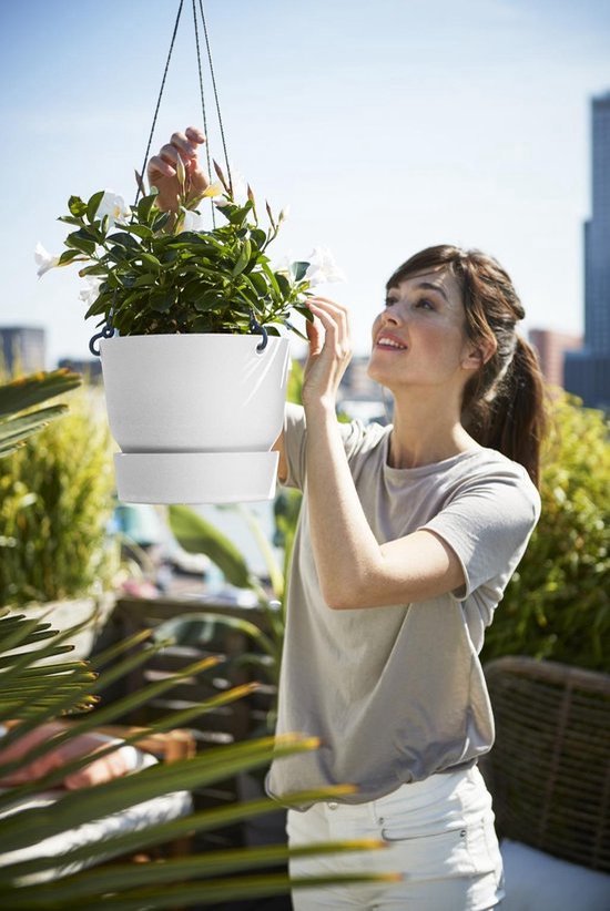 Vrouw met witte broek en grijze t-shirt verzorgt een witte hangpot met witte bloemen.