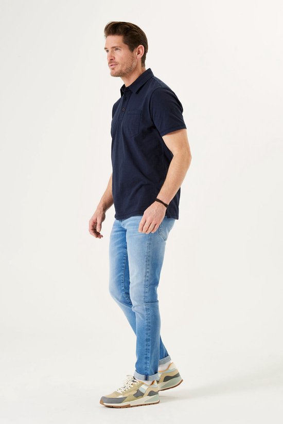 Man met donkerblauwe polo en lichtblauwe jeans, staand opzij.
