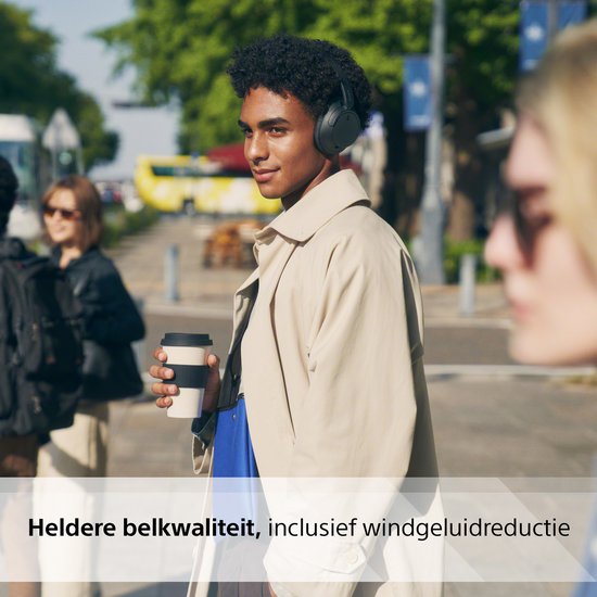 Vrouw met zwarte Sony koptelefoon, kofﬁe in hand, buiten op straat.