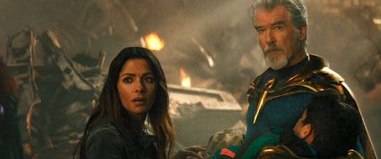 Pierce Brosnan als Dr. Fate, in een blauw kostuum met gouden accenten, die een gewonde jongen vasthoudt.