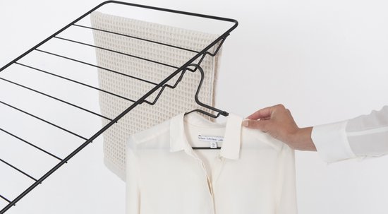 Een vrouw met een witte blouse en beige broek hangt een wit overhemd aan een zwarte kledinghanger, naast een vaalwit geblokt linnen doek op het droogrek.