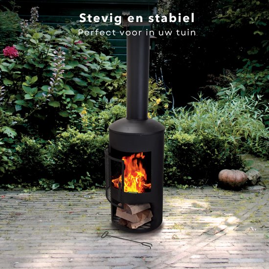 Zwarte, cilindervormige tuinhaard met brandend vuur en houtblokken, in een tuin met stenen en groen.