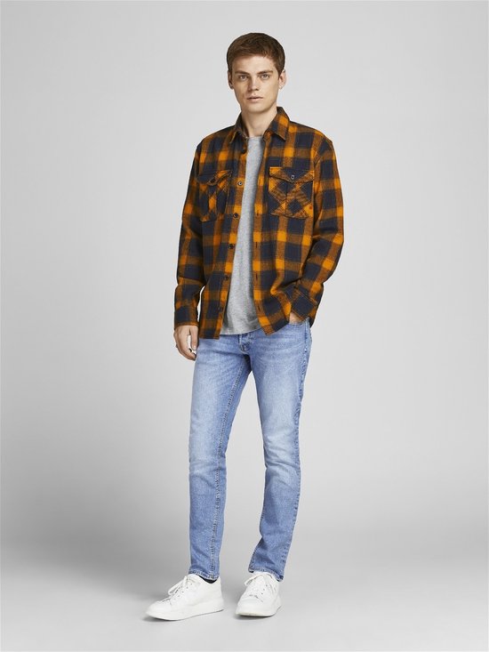 Man met een oranje met zwart geblokt overhemd en lichtblauwe skinny jeans, staand.