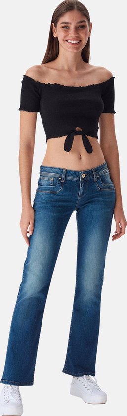 Model met een zwarte off-shoulder top met strikdetail en blauwe bootcut jeans met zichtbare knopen.