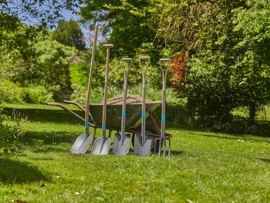 Vier tuingereedschappen: een schop, een spit, een plant- en plantguts, en een schoffel, staand voor een kruiwagen vol zand in een tuin.