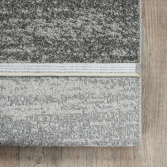 Detailopname van een grijs vloerkleed met een gedessineerde bovenkant en een effen, geweven onderkant op een houten vloer.