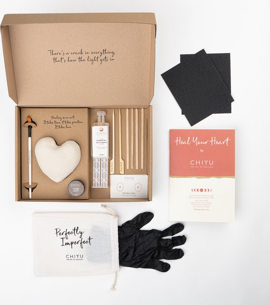 Open Kintsugi 'Heal Your Heart' doos met een witte hartvorm, reparatiebenodigdheden en een 'Heal Your Heart' boekje. De binnenkant van de doos heeft de tekst 'There's a crack in everything, that's how the light gets in'.