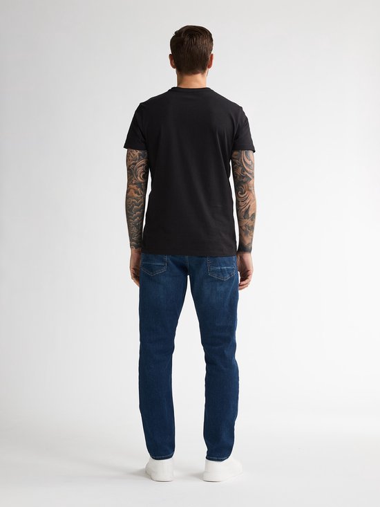 Man met tatoeages draagt een zwarte t-shirt en donkerblauwe jeans. De jeans hebben een normale pasvorm en witte sneakers.