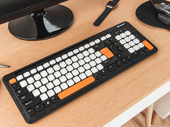 Zwart QWERTY-toetsenbord met witte en oranje toetsen, op een houten bureau naast een monitor.