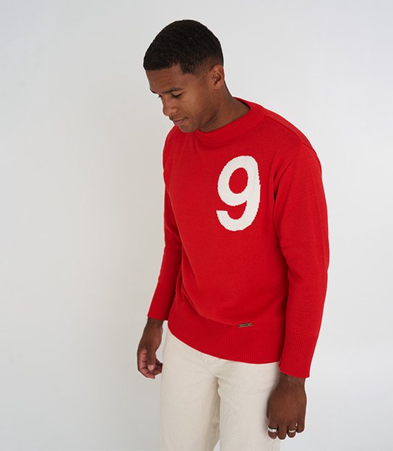 Man, kijkend naar beneden, in een rode trui met witte 9, gedragen met een beige broek.