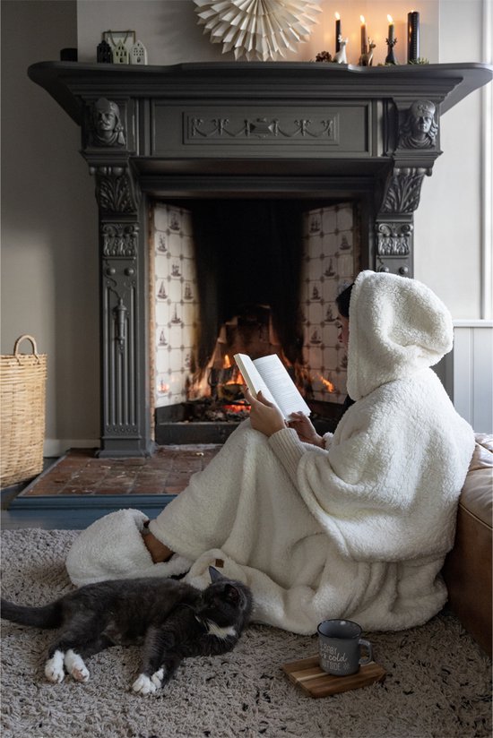 Een persoon in een witte teddy hoodie deken leest een boek bij een open haard, met een kat op de grond.