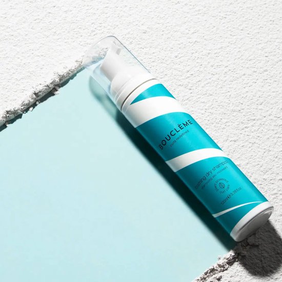 Fles met teal- en witte spiraalvormige strepen voor droogshampoo, liggend op een lichtblauwe en grijze ondergrond.