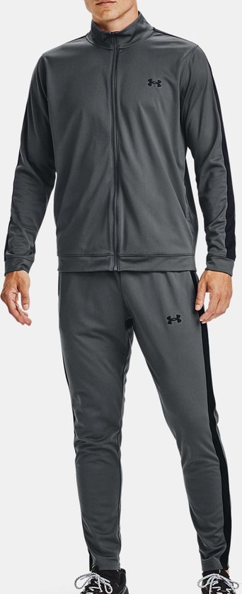 Man met een grijs Under Armour trainingspak met zwarte strepen op de mouwen en zijkanten van de broek. Zwarte logo's.