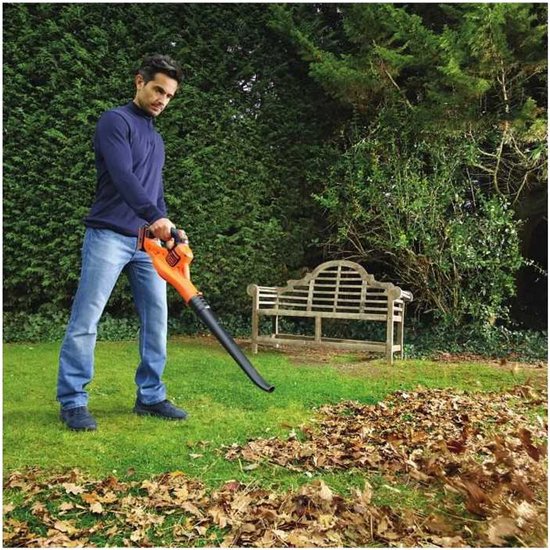 Man in blauwe trui en spijkerbroek gebruikt oranje bladblazer in tuin met herfstbladeren.