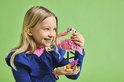 Een lachend meisje met blond haar, in een blauwe jurk, houdt een roze LEGO flamingo vast met bloemen.